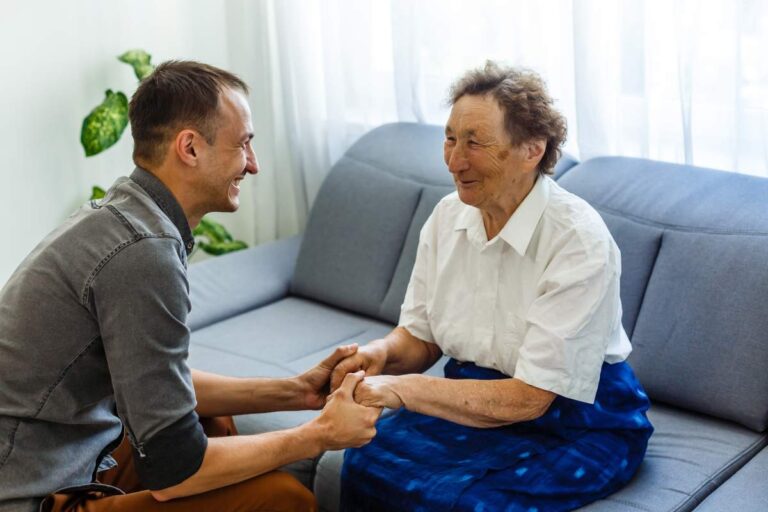 An older adult and a caregiver share a moment of conversation at a kitchen table with coffee cups.